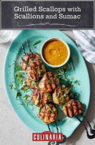 Two grilled scallop kabobs on a blue oval platter with a dish of dipping sauce on the side.