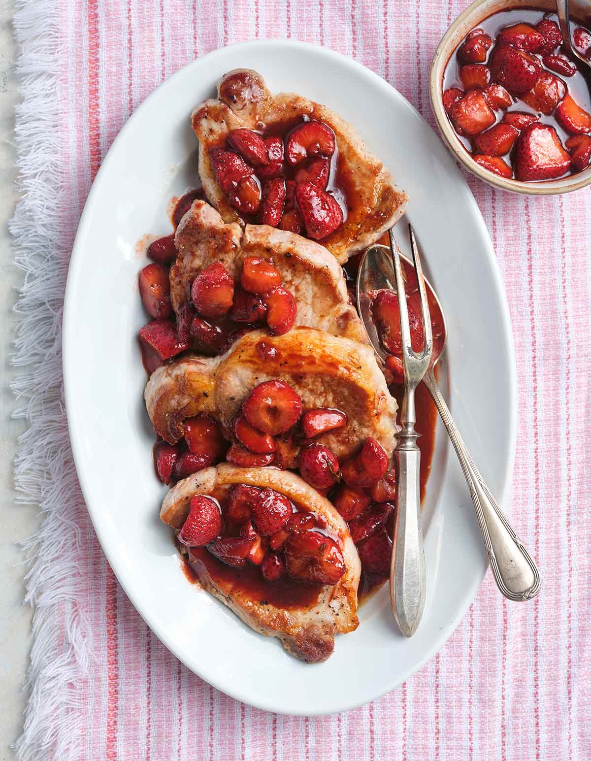 Four pork chops with strawberry balsamic glaze on top arranged on a white oval platter with serving utensils and a bowl of strawberry glaze on the side.