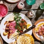 A platter of grilled boneless chicken thighs garnished with pickled red onions, mint, lemon and pita sides.