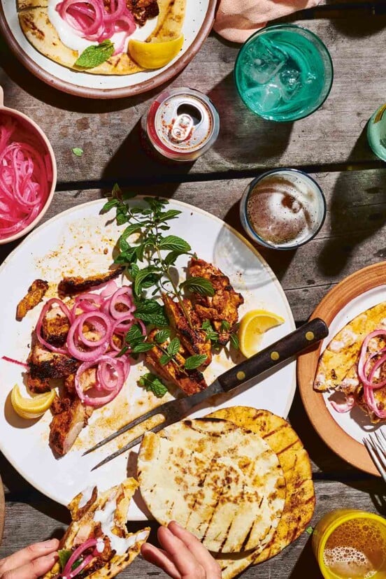 A platter of grilled boneless chicken thighs topped with pickled red onion, mint, lemon, and pita on the side.