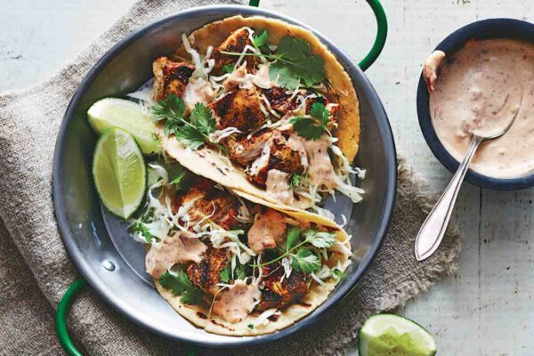 Two ancho fish tacos in a metal dish with lime wedges and chipotle mayo on the side.