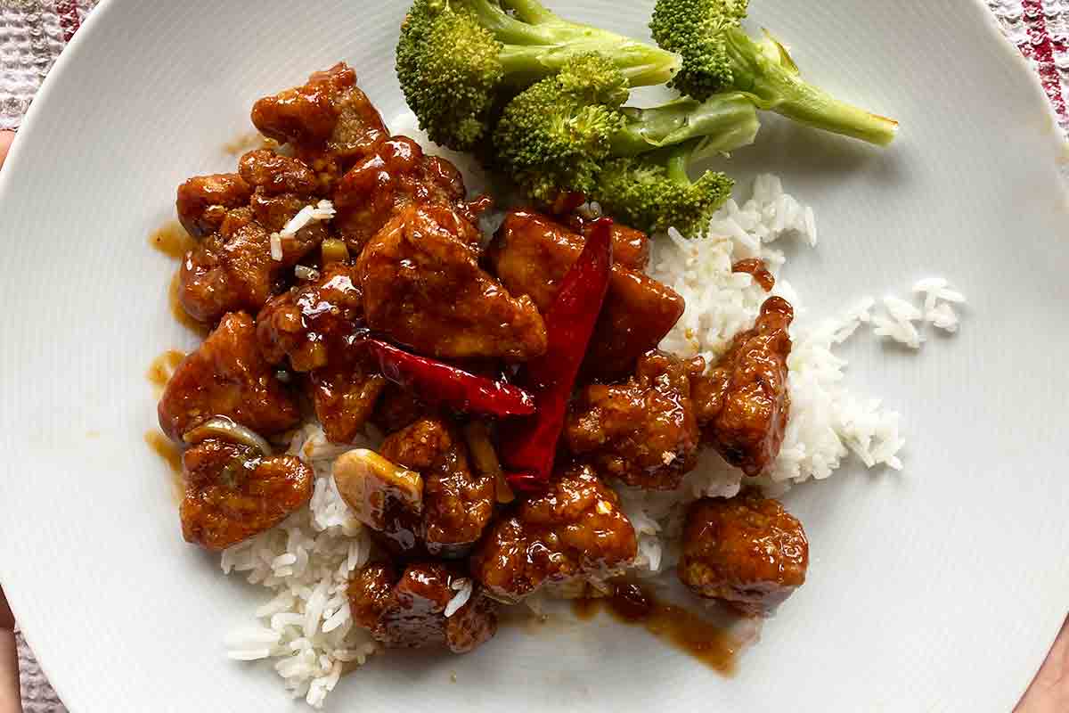 A white plate with authentic General Tso's chicken on top of rice with steamed broccoli on the side.