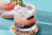 Two short glasses of blueberry prosecco spritz on coasters with thyme sprigs for garnish.