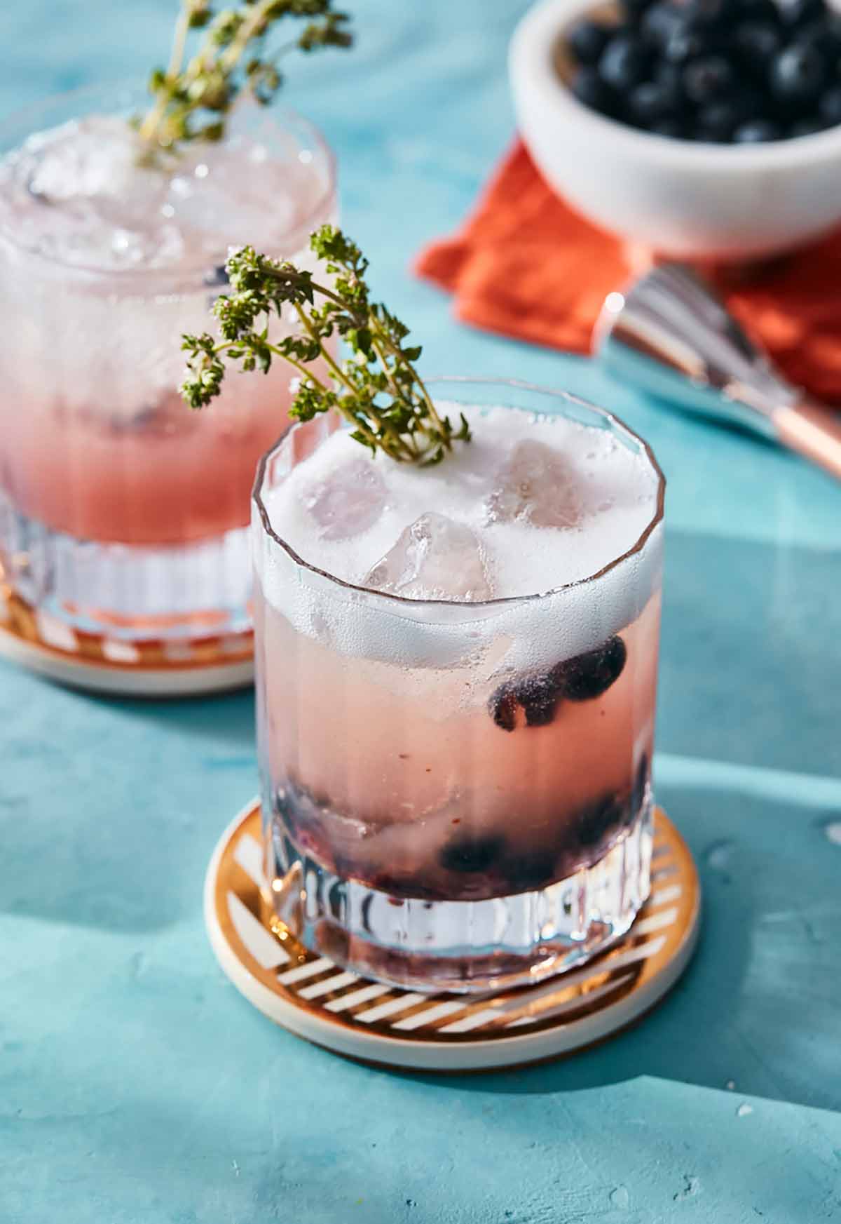 Two short glasses of blueberry prosecco spritz on coasters with thyme sprigs for garnish.