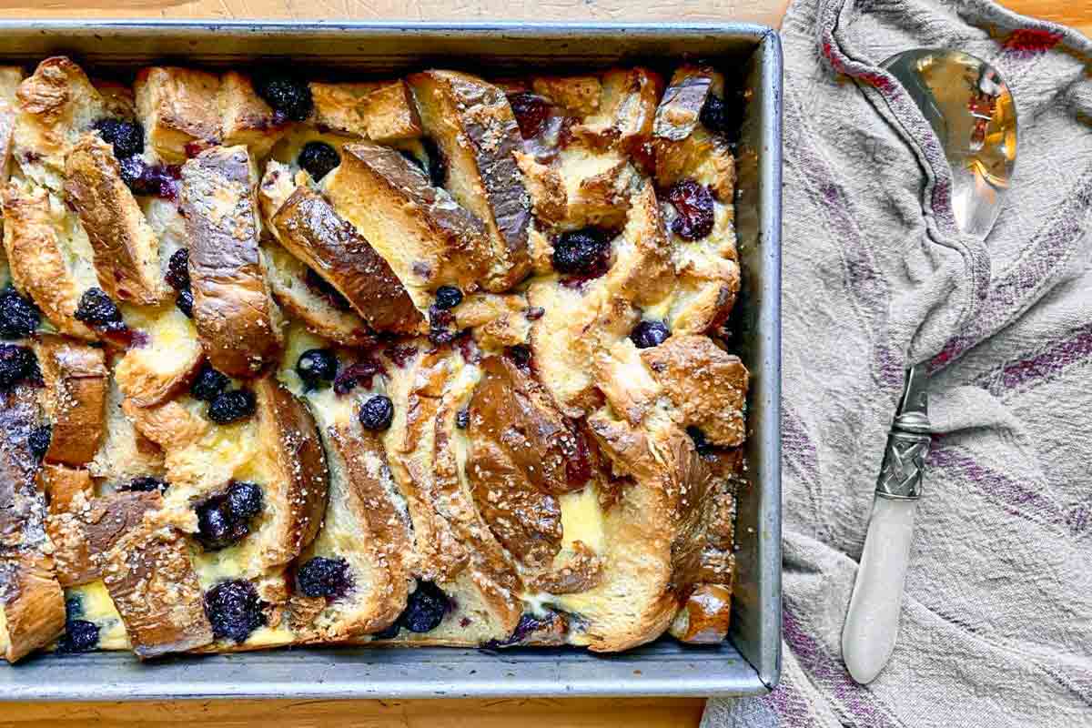 A rectangular pan of blueberry brioche French toast casserole next to a kitchen towel and fancy serving spoon.