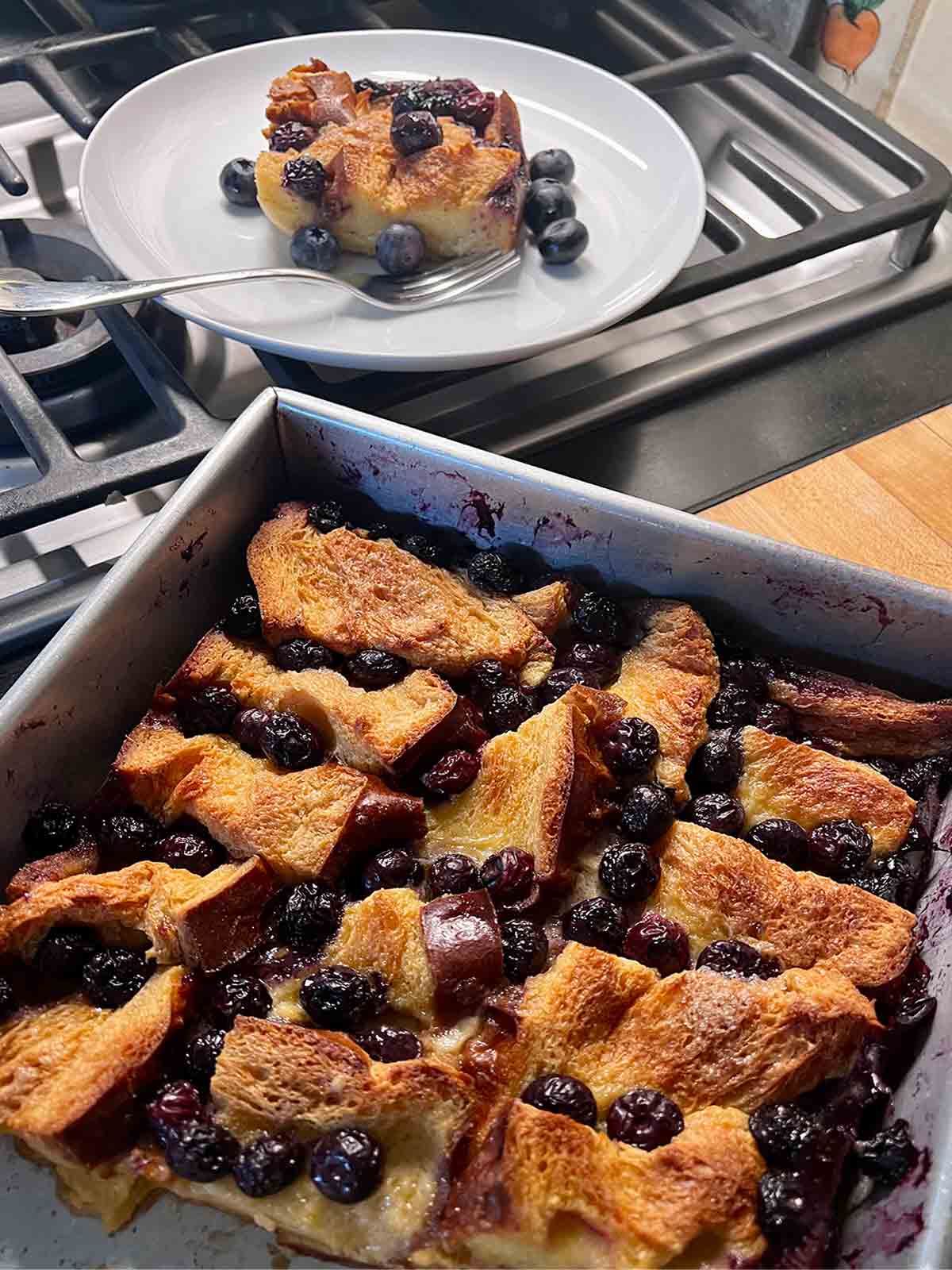 Half of a rectangular pan of blueberry brioche French toast casserole next to a gas range with a serving of the casserole on a white plate.