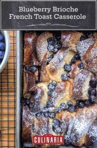A pan of blueberry brioche French toast casserole on a wire rack with a bowl of blueberries next to it.