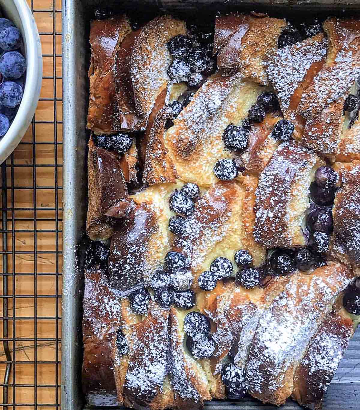 A pan of blueberry brioche French toast casserole on a wire rack with a bowl of blueberries next to it.