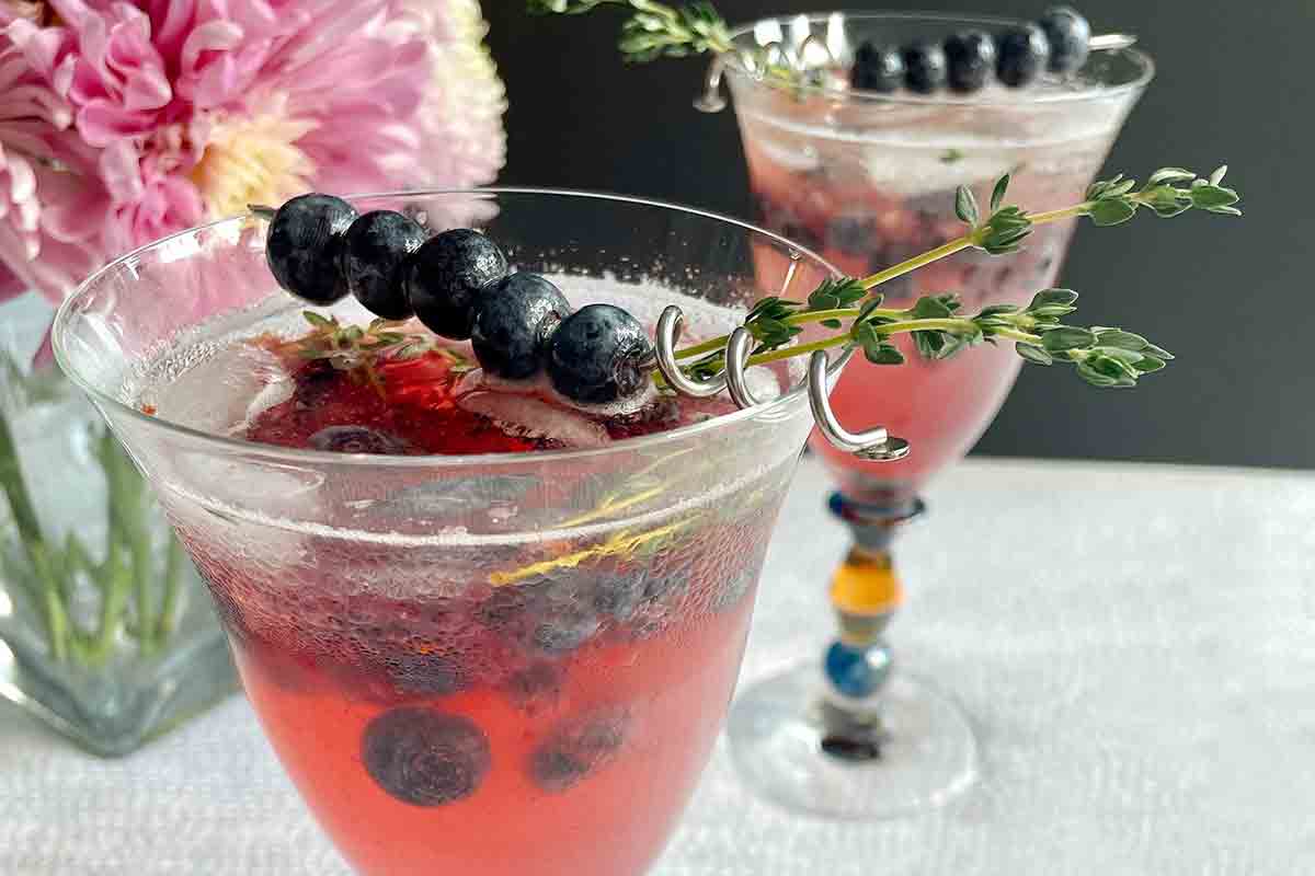 Two glasses of blueberry prosecco spritz with a skewer of blueberries and a sprig of thyme on top.