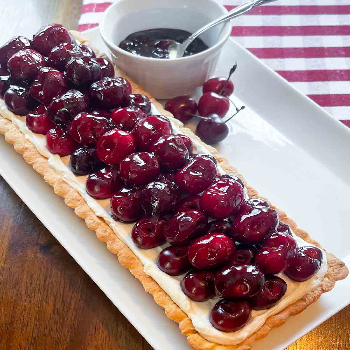 A fresh cherry tart with cream cheese filling on a white rectangular platter with a bowl of jam and a few fresh cherries on the side.