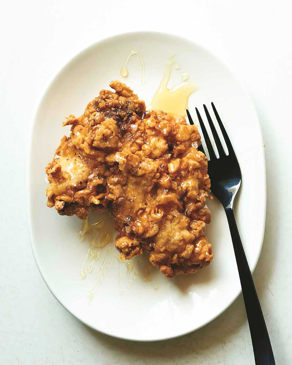 A fried boneless chicken thigh drizzle with honey on a white plate with a fork next to it.