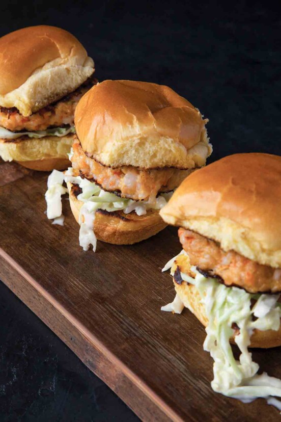 Three fried shrimp sliders with coleslaw and toasted Hawaiian rolls on a wooden board.