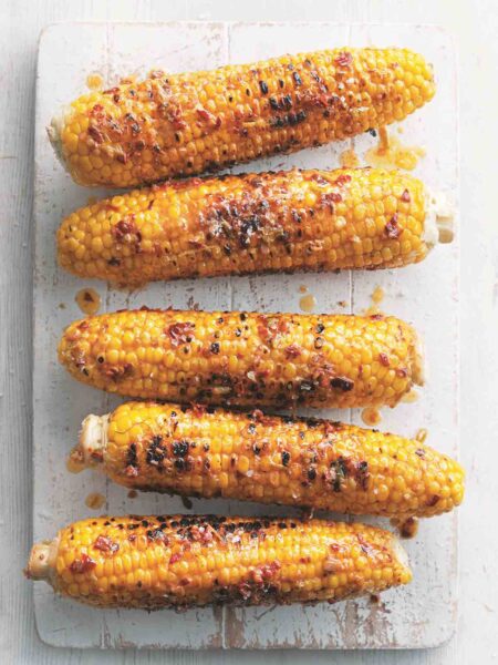 Five ears of grilled corn on the cob with chipotle butter on a white wooden board.
