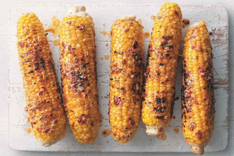 Five ears of grilled corn on the cob with chipotle butter on a white wooden board.