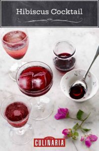 Four glasses filled with hibiscus cocktail with a bowl and spoon on the side and a few hibiscus flowers lying on the counter.