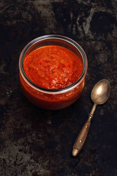 A jar of red homemade sriracha hot sauce on a black background. A spoon nearby.