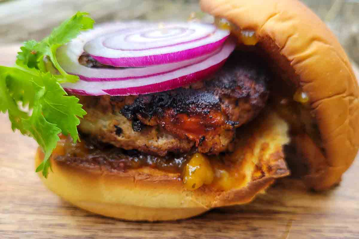 An Indian spiced chicken burger on a bun, topped with red onion slices and cilantro leaves.
