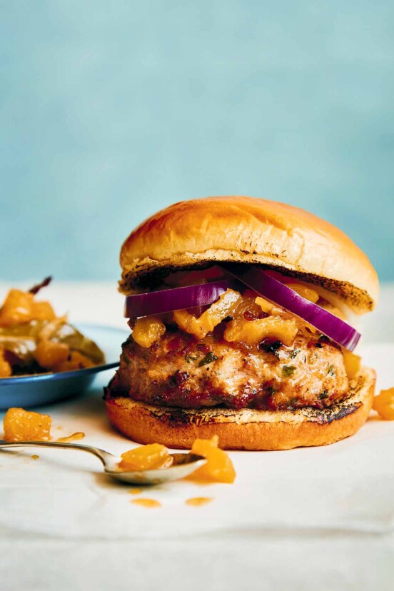 An Indian spiced chicken burger, with chutney and red onions on top and a bowl of chutney on the side.