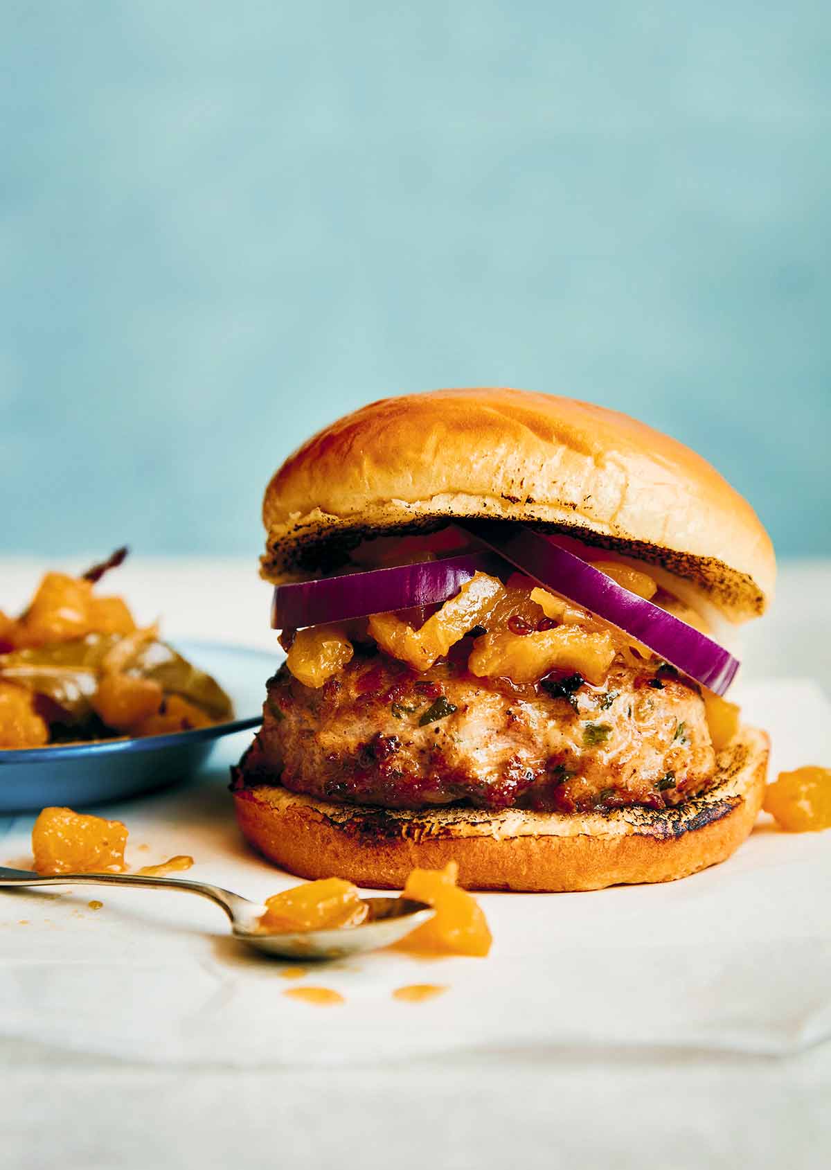 An Indian spiced chicken burger, with chutney and red onions on top and a bowl of chutney on the side.