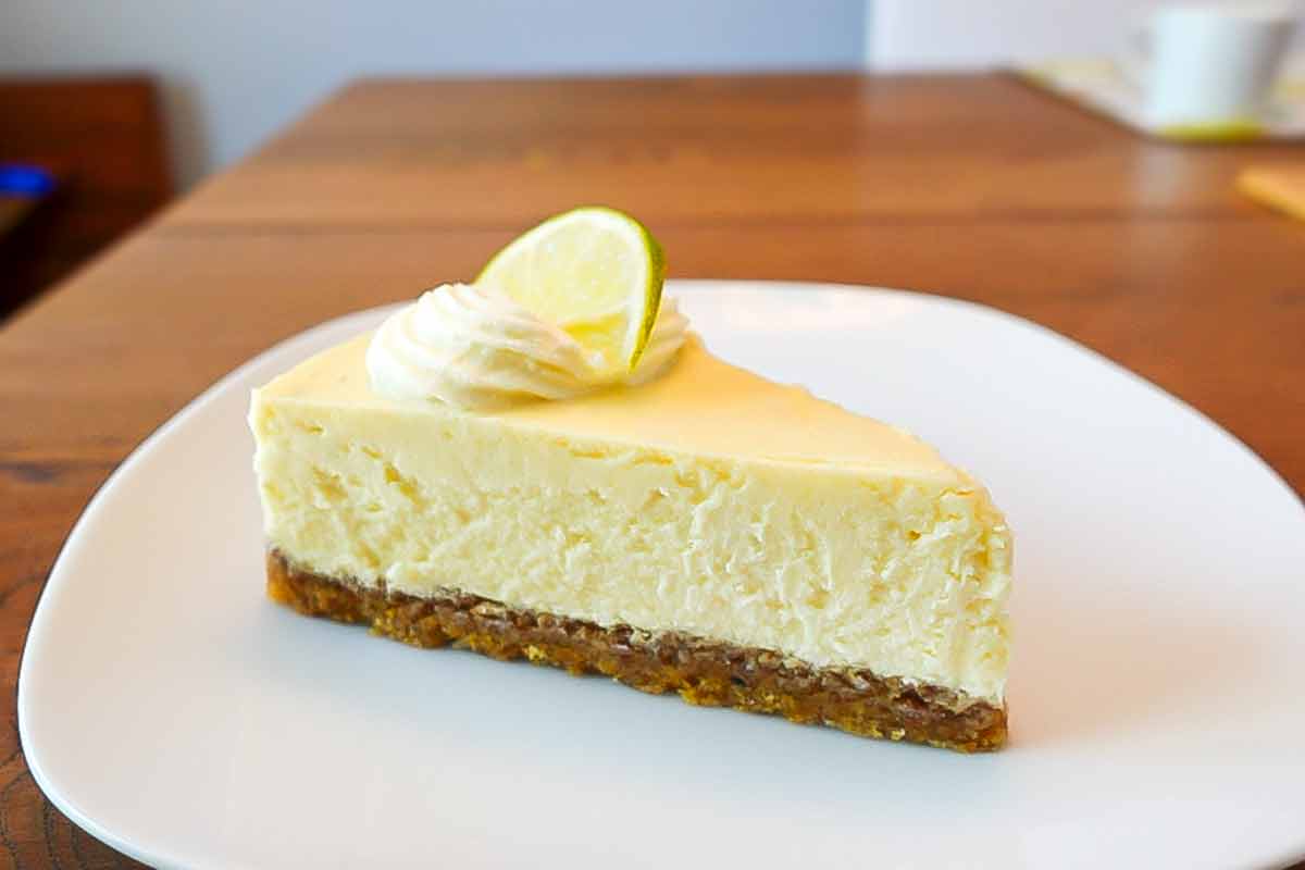 A slice of key lime cheesecake with pecan crust on a white plate, topped with a lime slice and a dollop of whipped cream.
