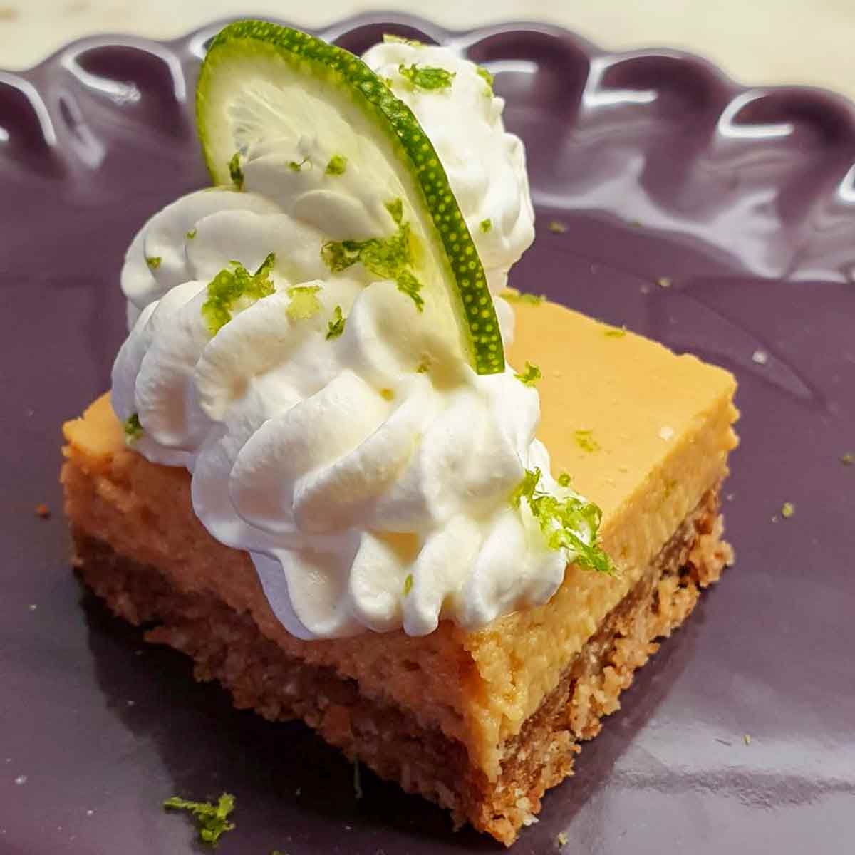 A square key lime pie bar with whipped cream and a lime slice on top on a purple plate.