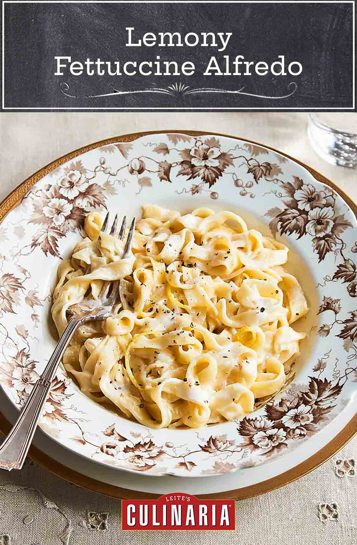 A patterned bowl filled with lemony fettuccine alfredo on a white and gold plate with a glass of water and dish of Parmesan on the side.