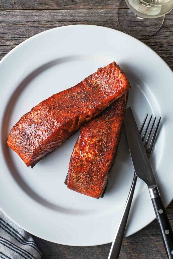 Two pieces of maple smoked grilled salmon on a white plate with a fork and knife on the side.