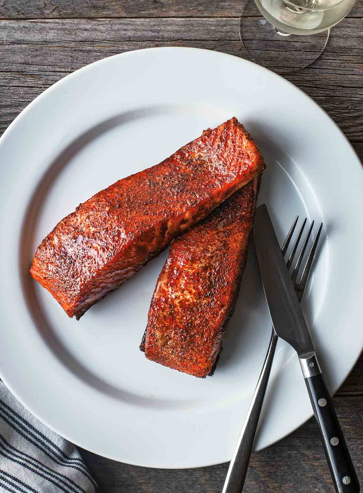 Two pieces of maple smoked grilled salmon on a white plate with a fork and knife on the side.