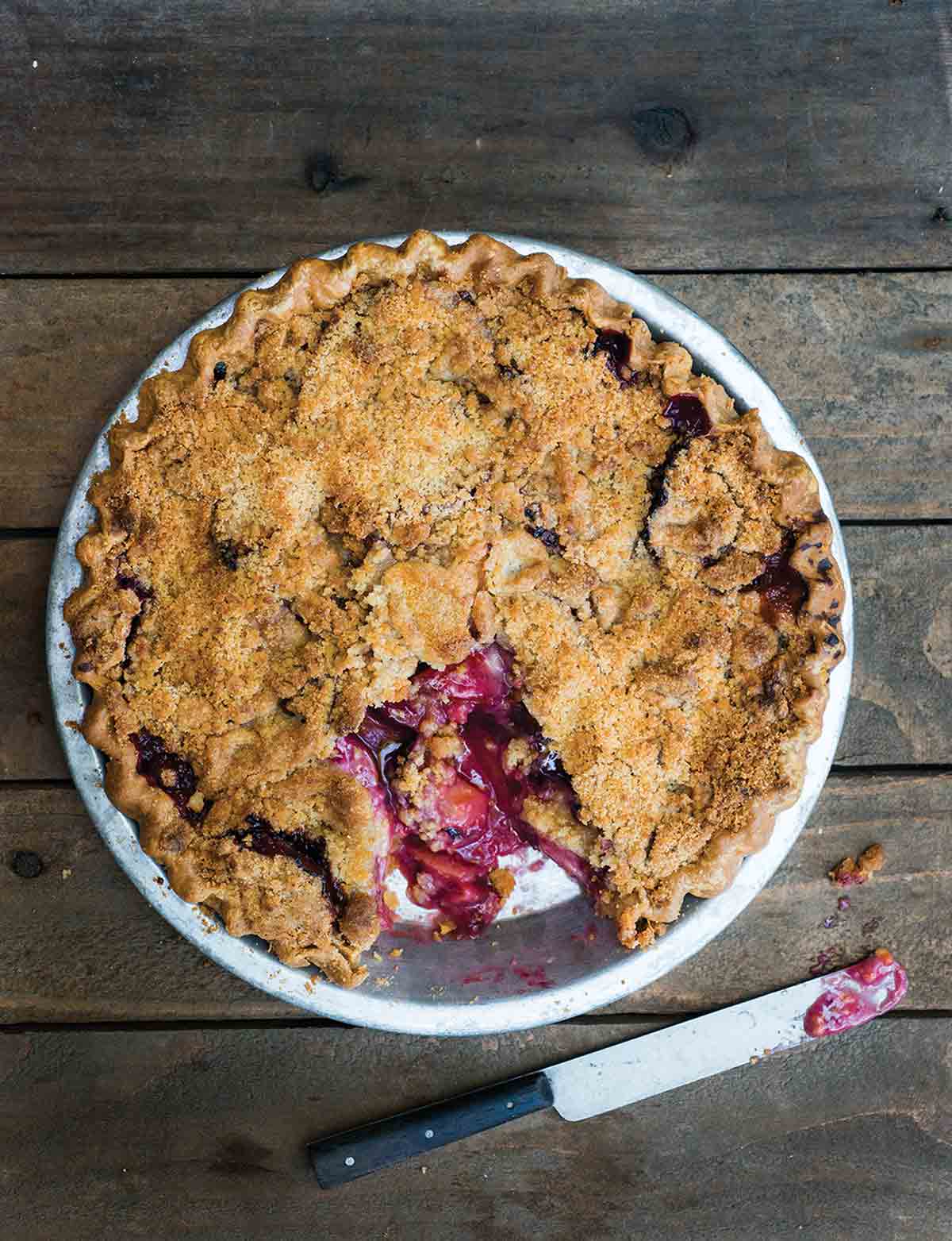 A baked plum crumble pie with a serving cut from it and a knife on the table beside it.