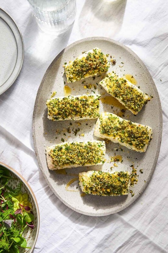Six fillets of quick baked fish with bread crumbs and herbs on top on an oval platter.