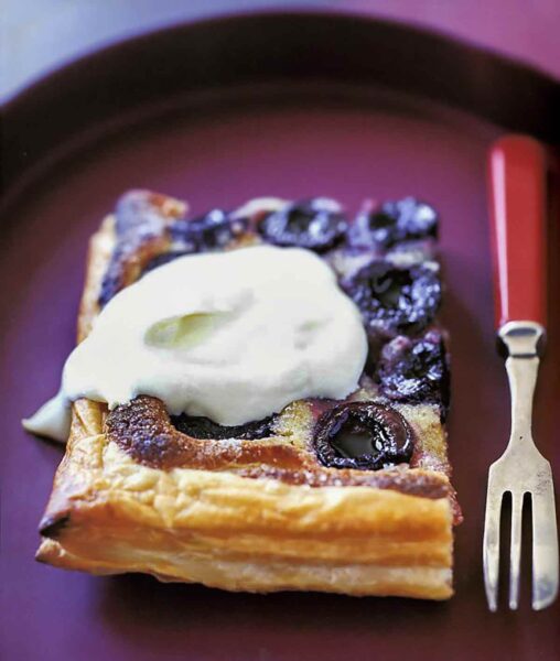 A square of rustic cherry puff pastry tart, topped with a dollop of whipped cream.