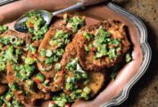 Several seared salt and pepper pork chops topped with a scallion and pepper mixture on a platter with a spoon resting on the side.