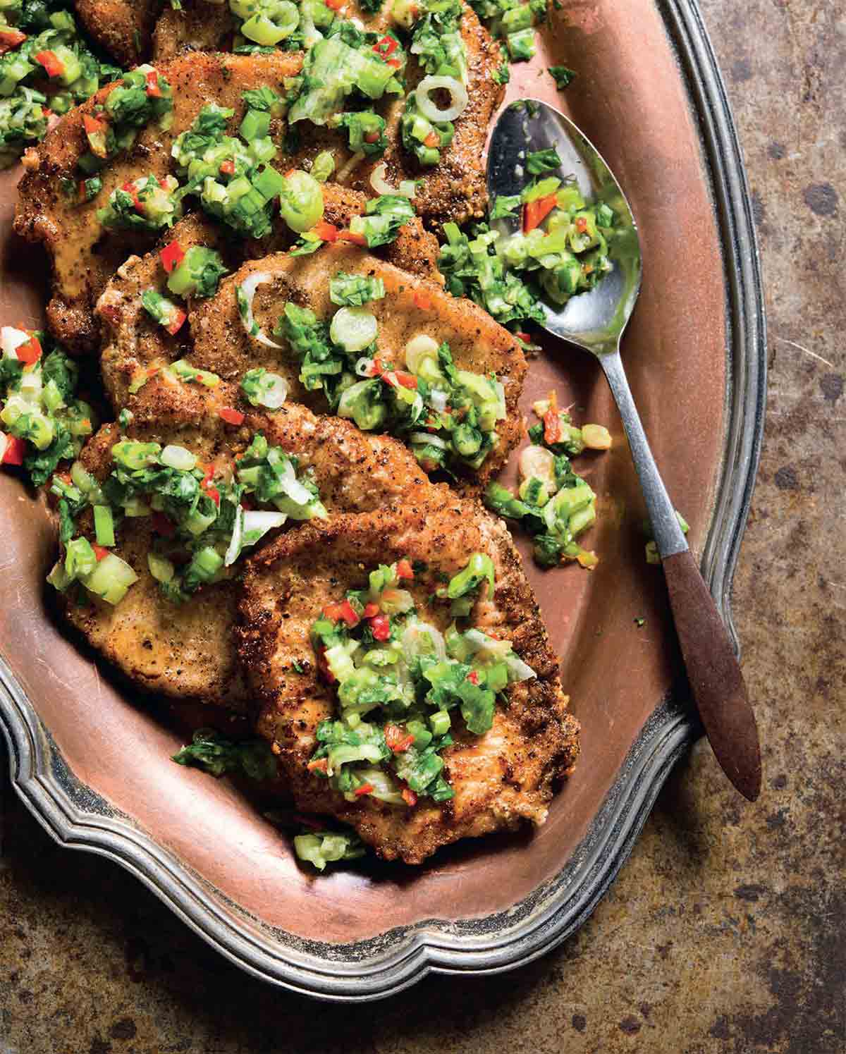 Several seared salt and pepper pork chops topped with a scallion and pepper mixture on a platter with a spoon resting on the side.