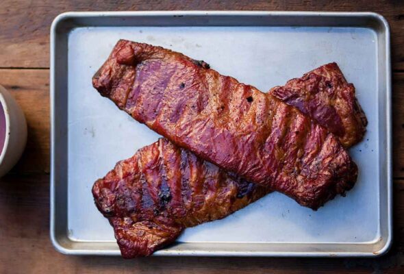 Two slabs of smoked ribs on a rimmed baking sheet with spicy bourbon bbq sauce in a dish on the side.
