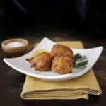 Three Spanish cod fritters, aka buñuelos de bacalao, on a square white plate with parsley on a yellow napkin and black tray