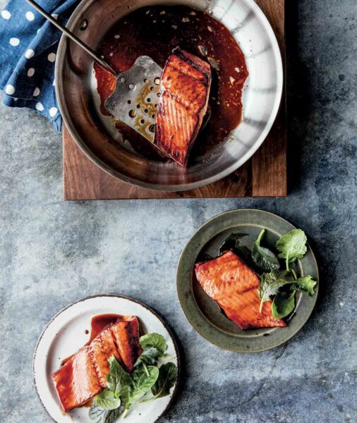A skillet on a wooden board with a piece of salmon teriyaki in it and a metal spatula and two plates with salmon teriyaki and greens on the side.