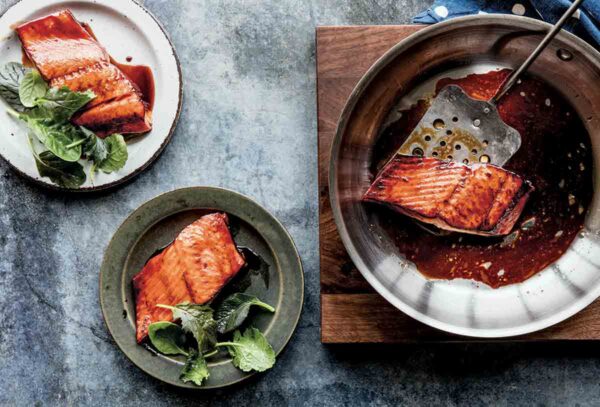 A skillet on a wooden board with a piece of salmon teriyaki in it and a metal spatula and two plates with salmon teriyaki and greens on the side.