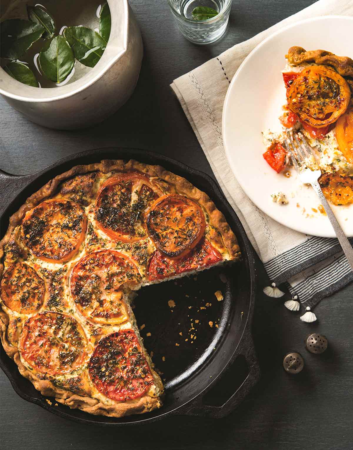 A cast-iron skillet filled with a tomato ricotta pie, one slice missing. Next to it, a white plate with a half eaten slice of pie.
