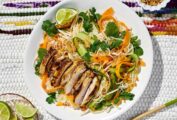 A white plate topped with vermicelli noodles, chicken, and raw vegetables with chopsticks and lime wedges on the side.