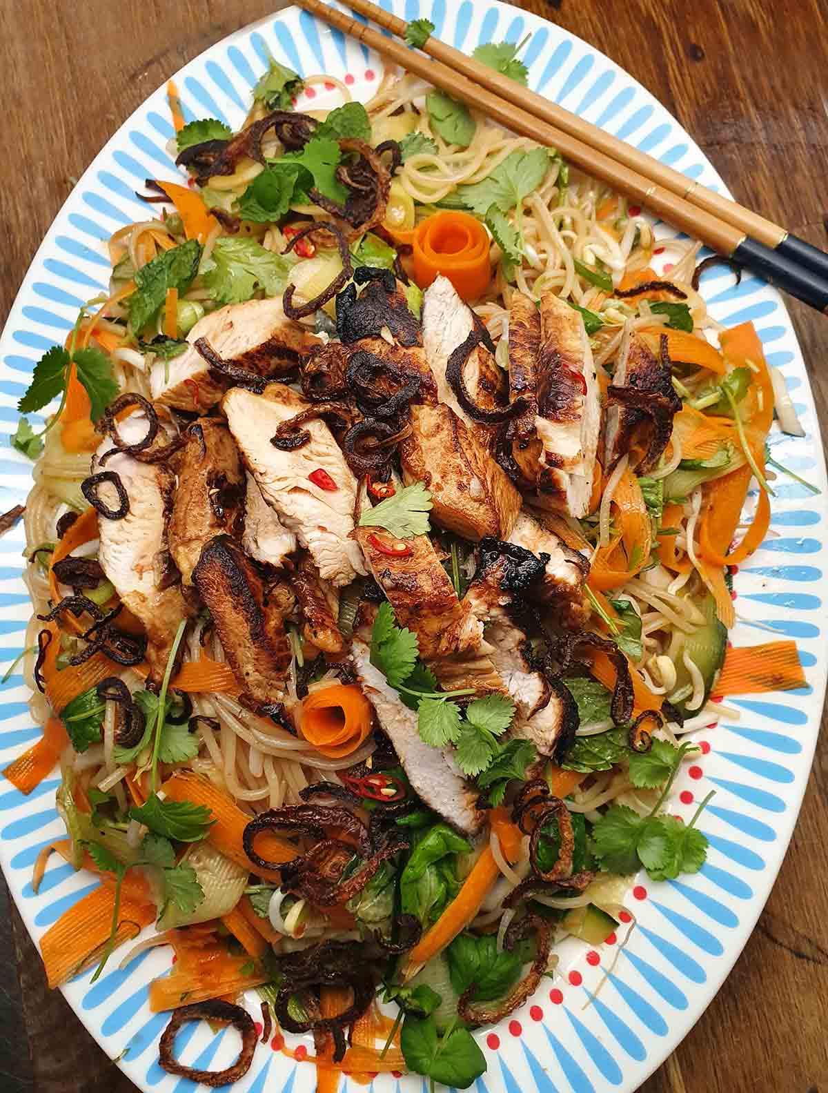 A blue and white oval platter with vermicelli noodles, chicken, raw vegetables and chopsticks on the side.