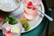 Two highball glasses filled with watermelon Moscow mule and topped with a mint sprig. A bowl of ice, watermelon and lime wedges on the side.