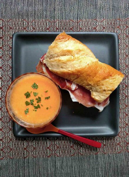 A square grey plate containing a bowl of cantaloup soup and a prosciutto mozzarella sandwich on a baguette.