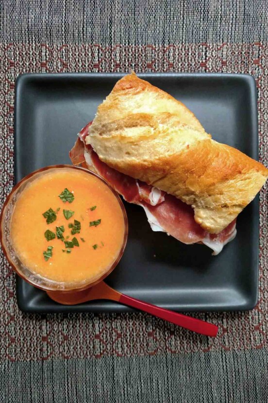 A square grey plate containing a bowl of cantaloup soup and a prosciutto mozzarella sandwich on a baguette.