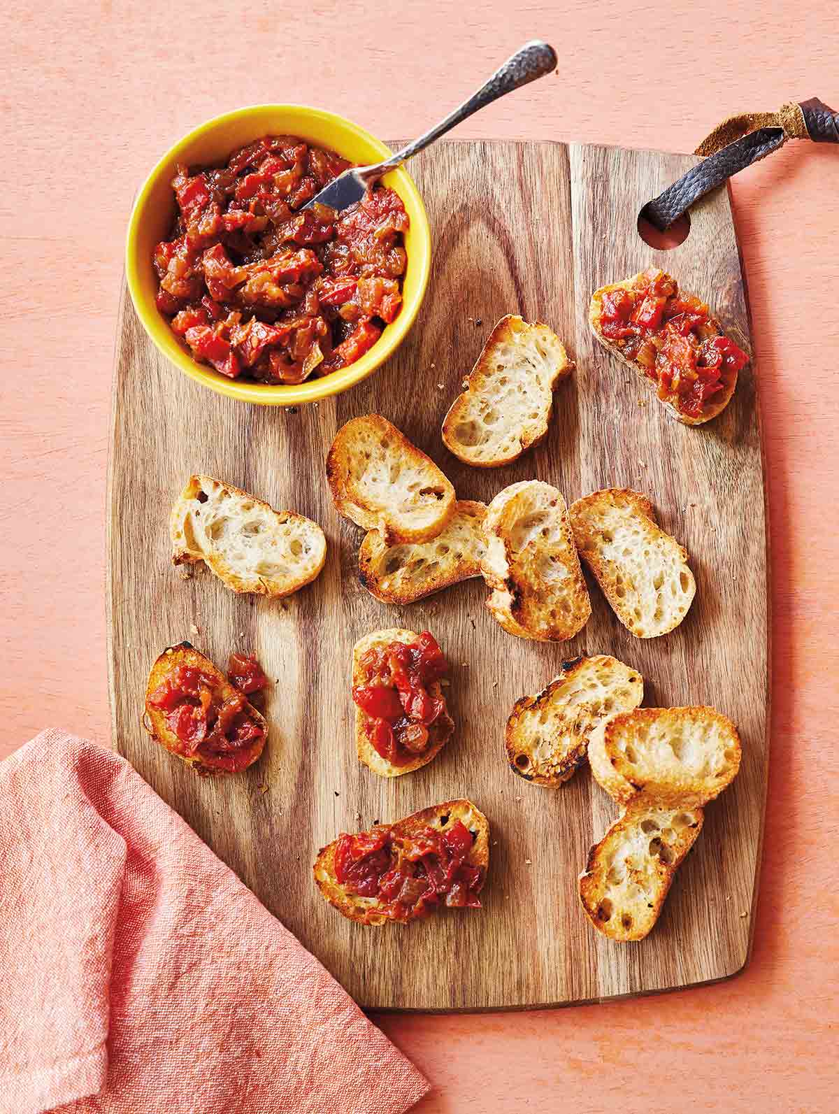A wooden cutting board with a bowl of caramelized onion and tomato jam and several toasted baguette slices on it.
