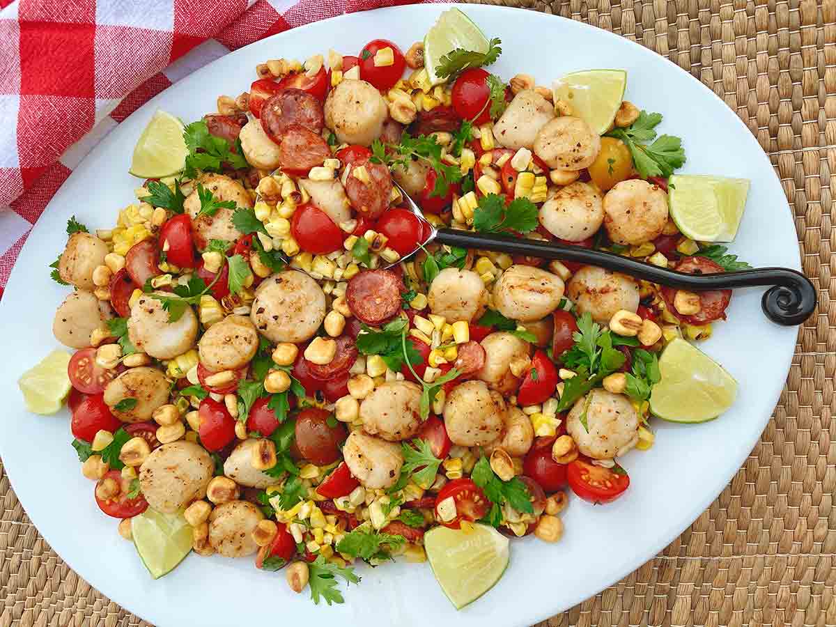 A white oval platter topped with charred corn and scallop salad, garnished with cilantro and lime wedges.