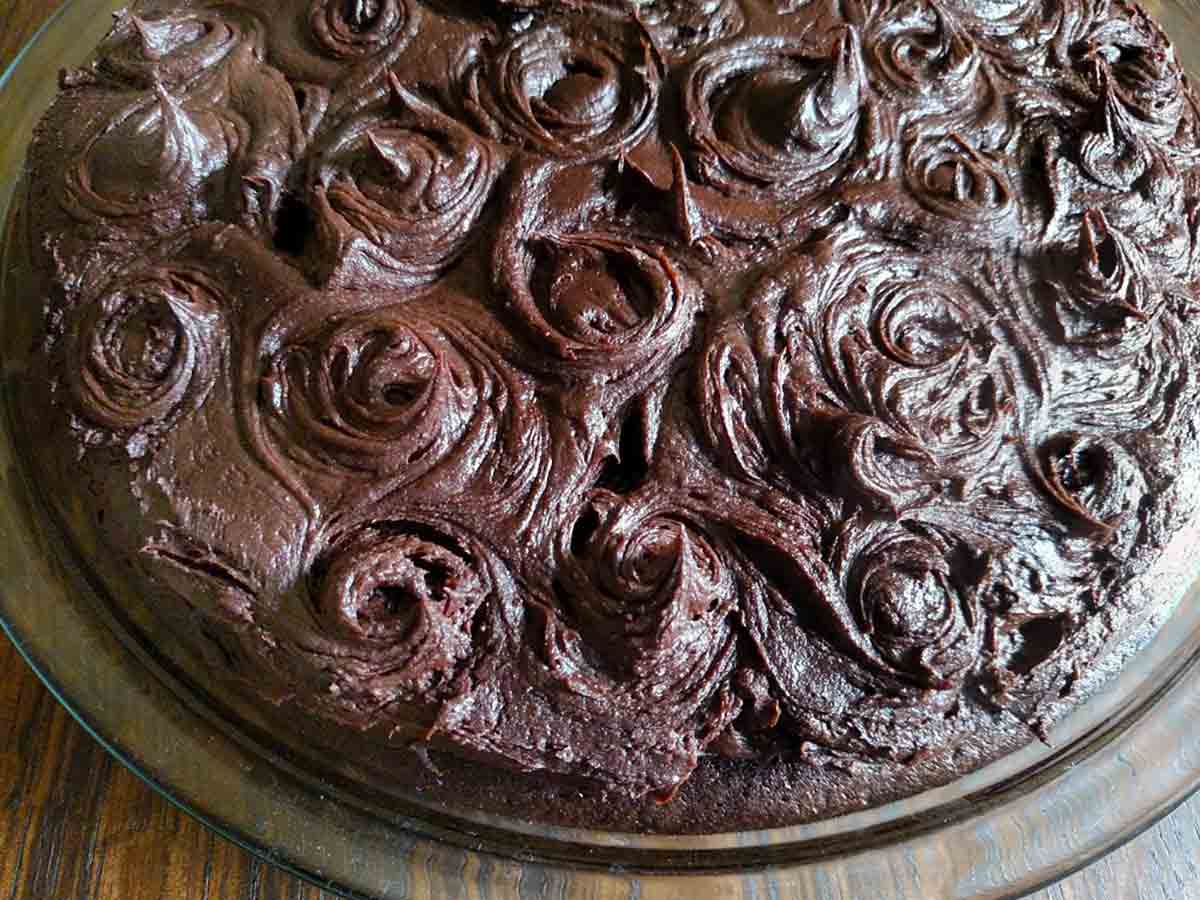 A chocolate wacky cake with chocolate frosting swirled on top on a glass serving plate.