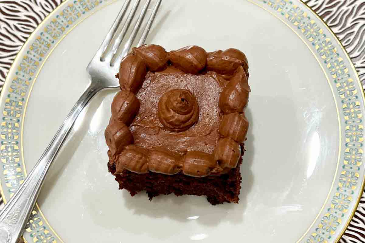 A square of chocolate zucchini cake with buttercream frosting on a china plate with a fork on the side.