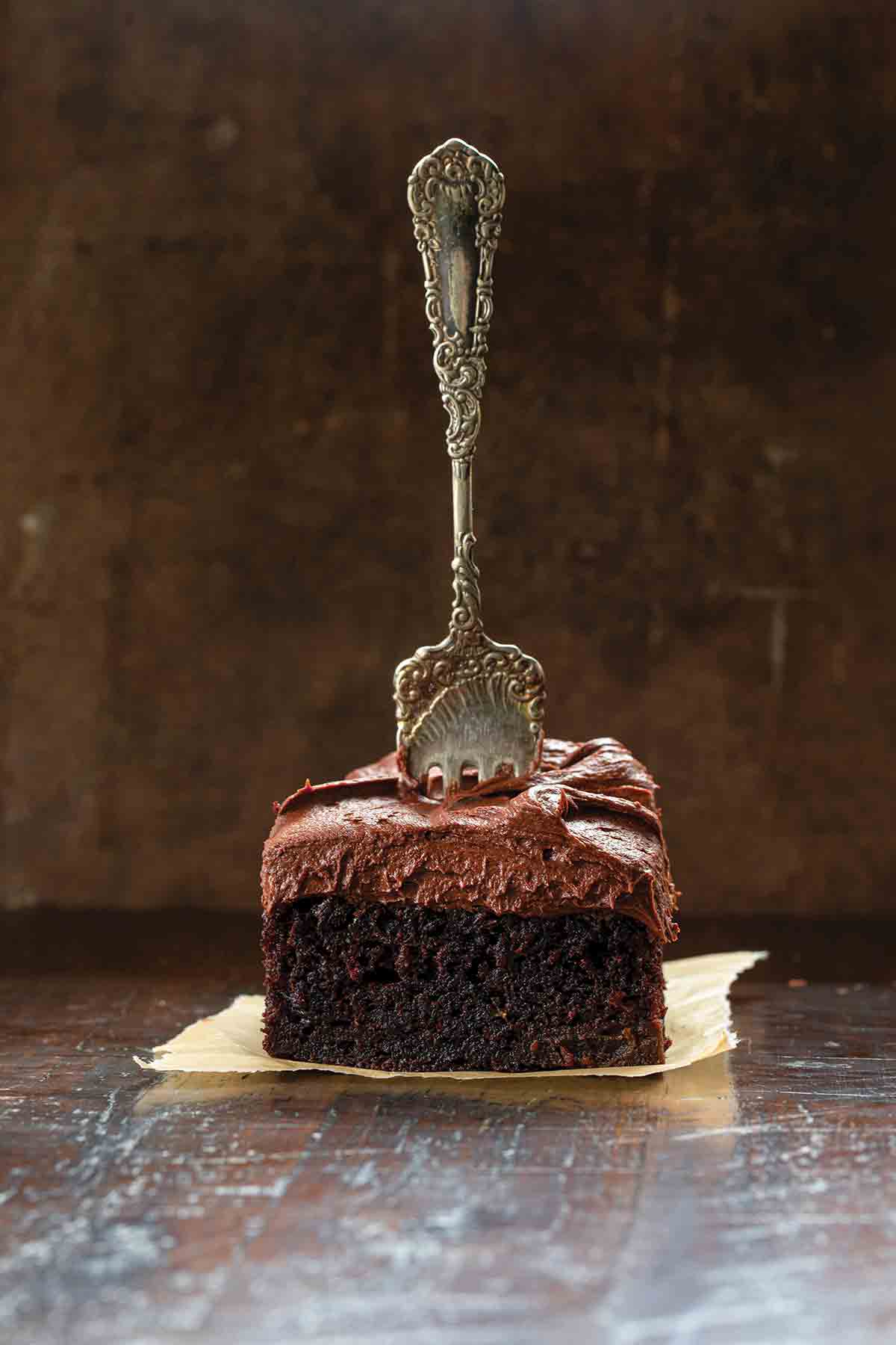 A square of chocolate zucchini cake with buttercream frosting on a square of parchment with a fork standing up in the center of the cake.