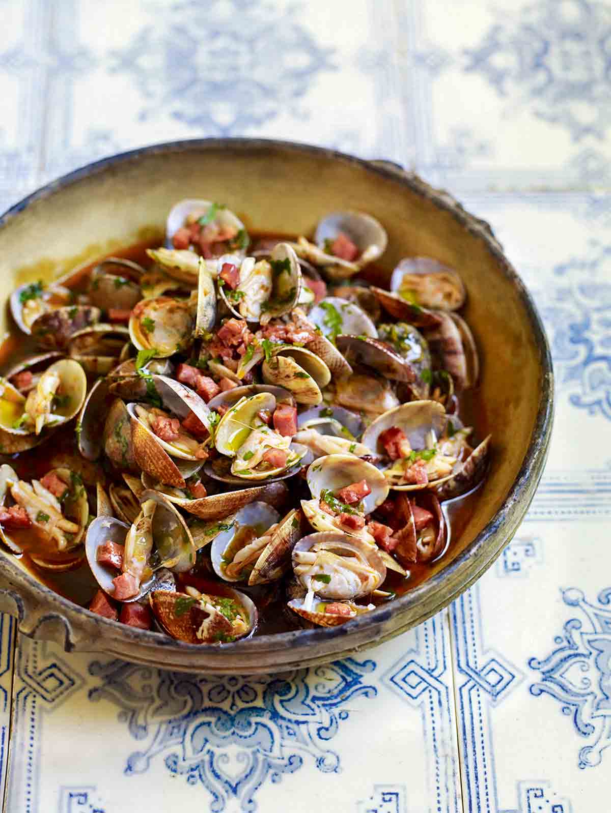 A bowl of clams with chouriço, garlic, and cilantro with a savory broth on top of blue and white tiles.