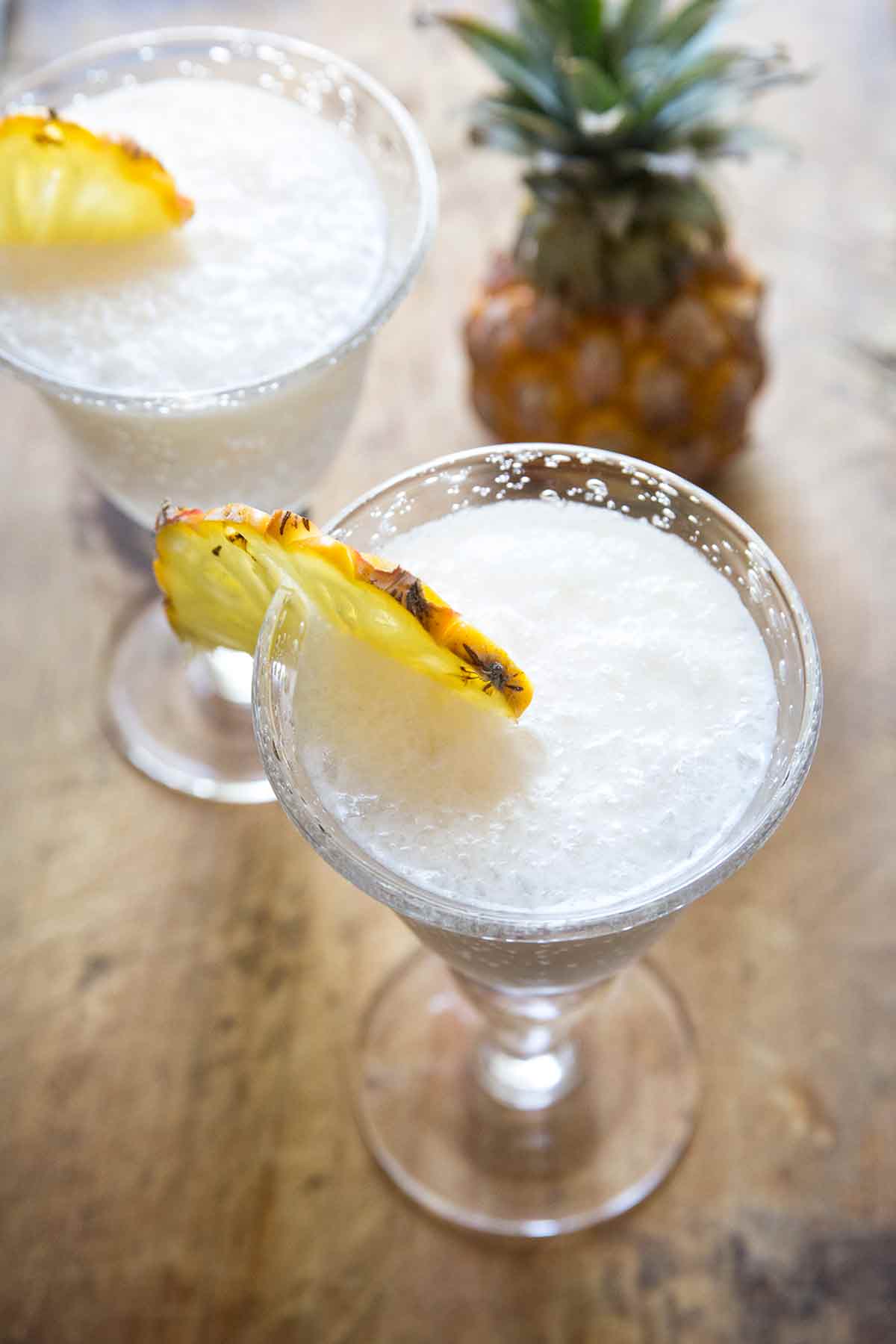 Two pina coladas, garnished with a pineapple wheel and a whole pineapple nearby.